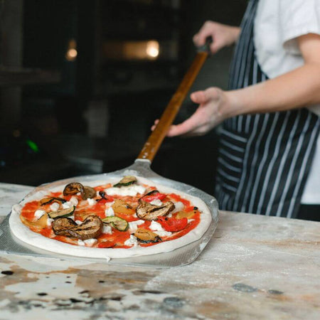 Person in a striped apron placing a pizza with mushrooms, mozzarella, and zucchini into a large commercial oven using a pizza peel.