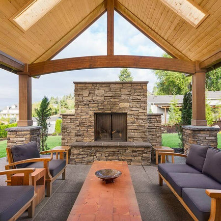 Outdoor seating area featuring lounge chairs and a table beneath a wooden roof. A stone fireplace is centrally located, surrounded by lush greenery and plants.