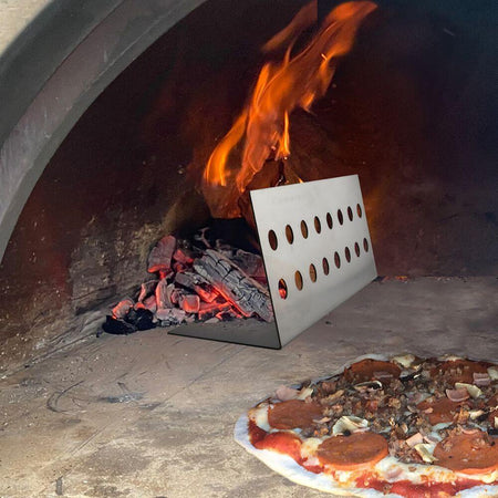 Pizza cooking in a wood-fired oven with flames in the background and a metal tool with holes placed near the embers.