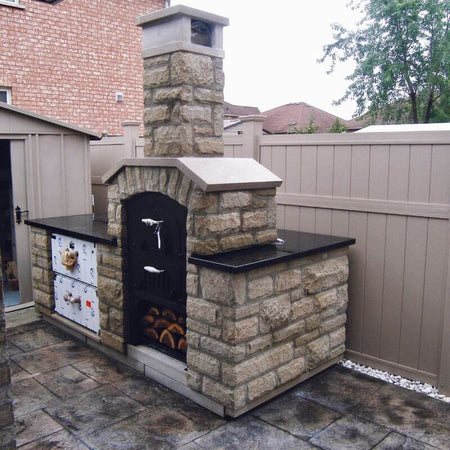 Standing point-of-view of a double-chamber wood-fired oven with fresh logs in its storage base, enclosed by a brick shell and countertop, set in a backyard.