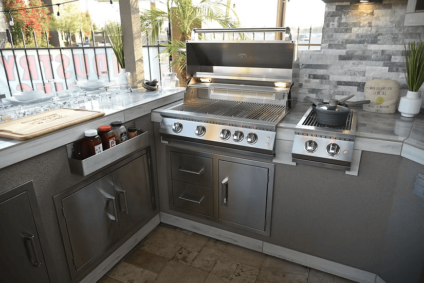 A modern outdoor kitchen with Kokomo Grills' Stainless Steel Two Drawer - One Door Combo (KO-ALPDC), gas grill, side burner, and utensils, all set against stone tile walls and a window.