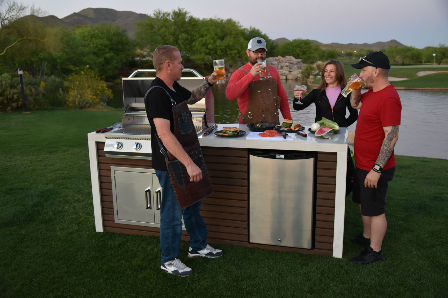 Four people gather around a Kokomo Grills Professional Shiplap Built-In BBQ Island Outdoor Kitchen with Waterfall Edge, enjoying drinks and food in a grassy yard with water and mountain views under a clear sky.