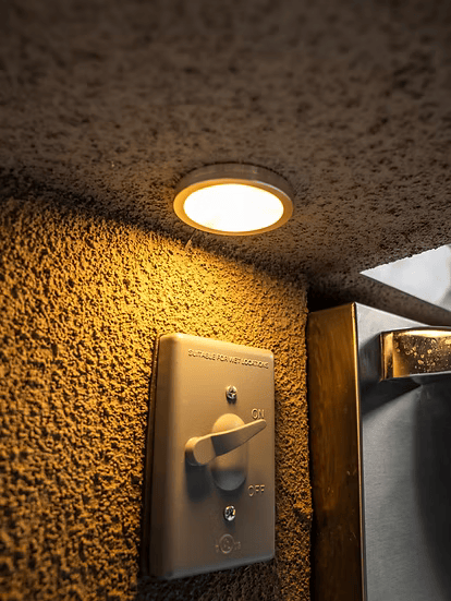A round recessed ceiling light glows above a textured wall with a beige switch labeled “ON” and “OFF,” next to the Kokomo Grills Mini Maui 6-Foot Kitchen Island by Kokomo Grills. The switch panel reads “SUITABLE FOR ART WORKUSE.”.