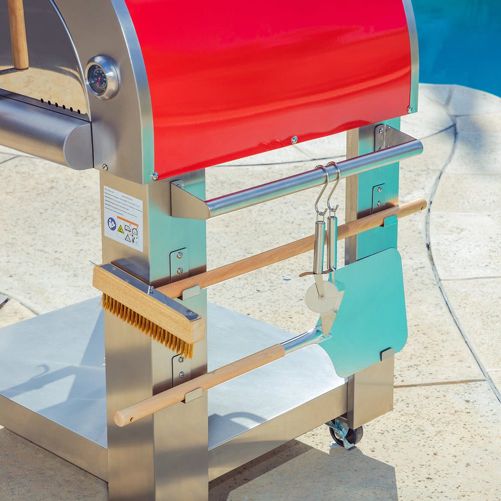 A close-up of the Empava Italian Red Outdoor Wood Fired Pizza Oven EMPV-PG06 with a stainless steel grill and barbecue tools, including brush, pizza peel, tongs, and cutter by the pool on a sunny day. Brand: Empava.