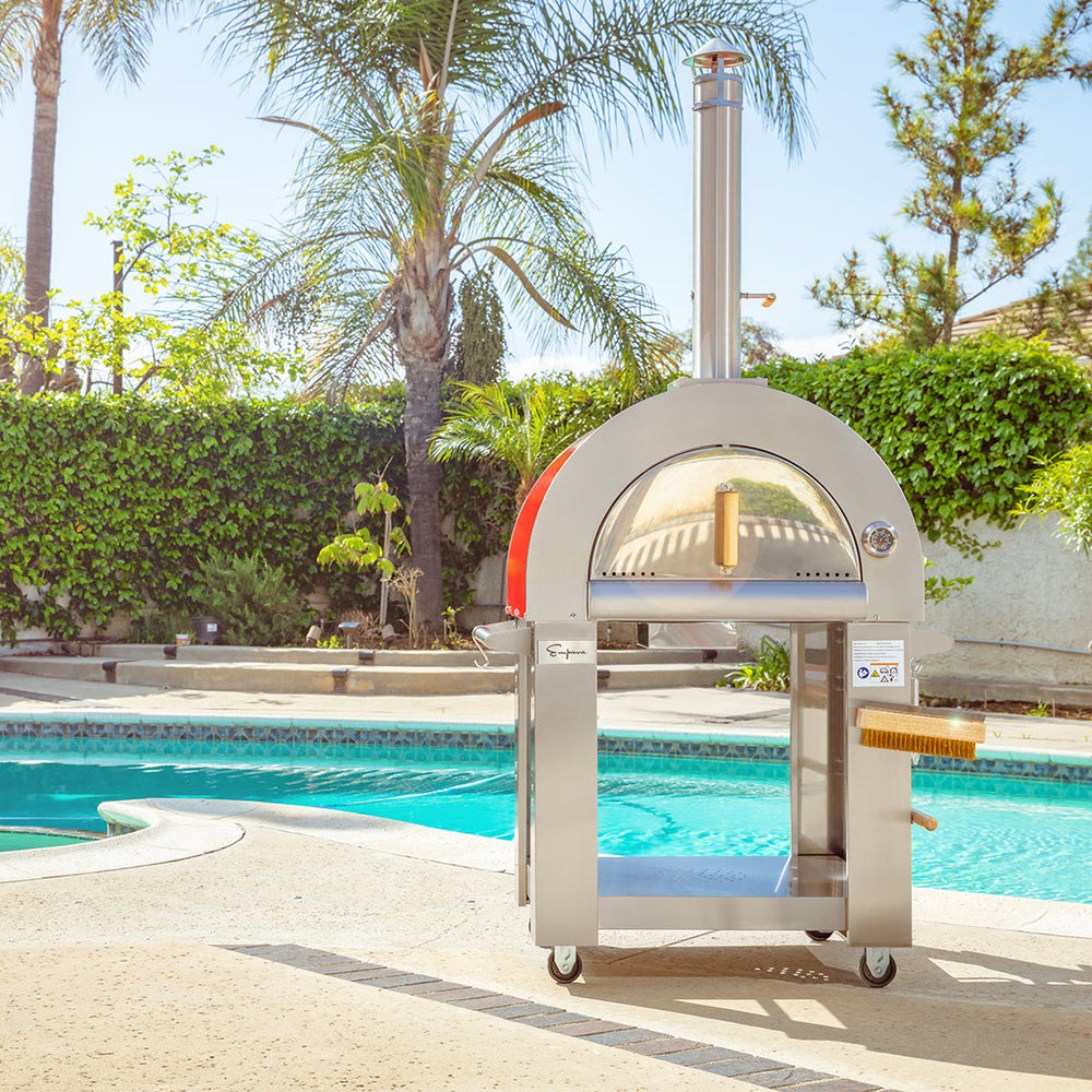 The Empava Italian Red Outdoor Wood Fired Pizza Oven EMPV-PG06 stands on a patio by a pool, surrounded by greenery and palm trees under the sun.