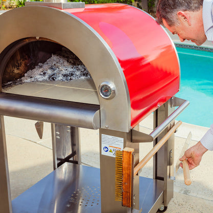 A man uses a wooden-handled brush to clean the Empava Italian Red Outdoor Wood Fired Pizza Oven EMPV-PG06, which features a stainless steel body and is placed beside a swimming pool.