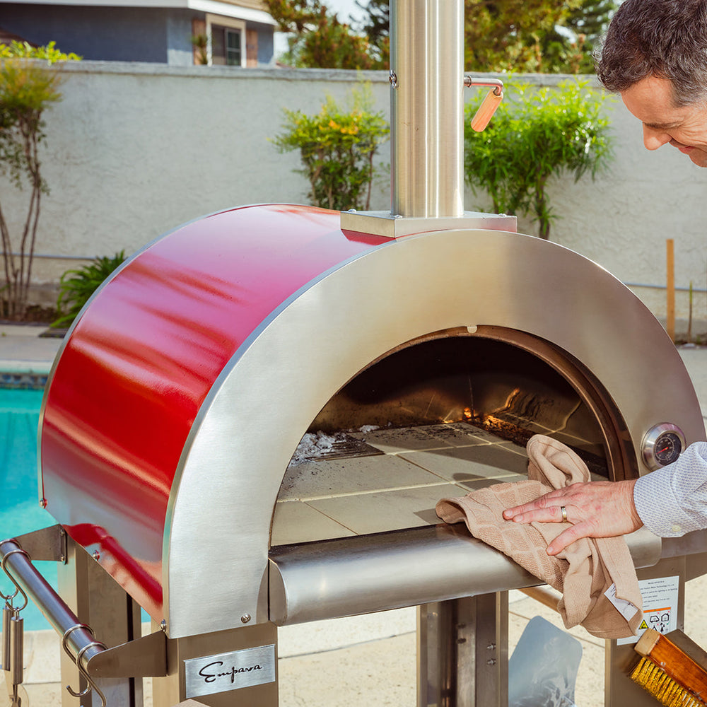 Someone cleans the stone surface inside an Empava Italian Red Outdoor Wood Fired Pizza Oven EMPV-PG06 beside a pool, with greenery and a house in the background.
