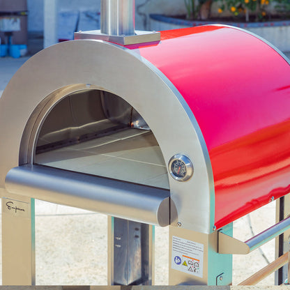 The Empava Italian Red Outdoor Wood Fired Pizza Oven EMPV-PG06 features a shiny curved top, stainless steel front, and built-in thermometer, and stands on a metal frame in a sunlit outdoor area.