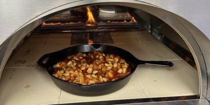 A Fire One Up PINNACOLO IBRIDO Hybrid Wood/Gas Outdoor Pizza Oven bakes chopped apples in sauce inside a cast iron skillet, with visible flames at the back.