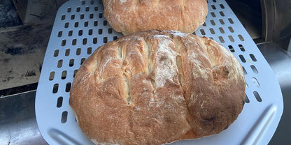 Two golden-brown rustic loaves with scored tops rest on a tray, fresh from the Fire One Up PINNACOLO IBRIDO (Hybrid) Wood/Gas Outdoor Pizza Oven. Their crisp crusts and light flour dusting show the quality of oven-fresh baking.