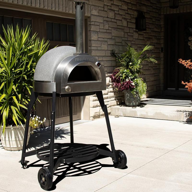 The Fire One Up PINNACOLO L’ARGILLA Gas Fired Outdoor Clay Pizza Oven with Accessories (PPO-8-08) stands on black metal legs with wheels on a sunlit patio near potted plants, in front of a stone house entrance.