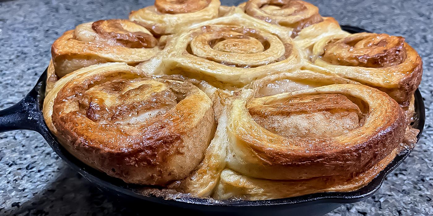 A close-up of golden brown cinnamon rolls arranged in a cast iron skillet, fresh from the Fire One Up PINNACOLO L’ARGILLA Gas Fired Outdoor Clay Pizza Oven (PPO-8-08), on a speckled countertop.
