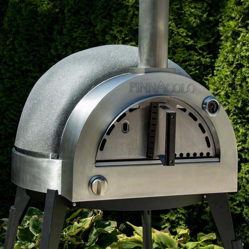 A Fire One Up PINNACOLO L’ARGILLA Gas Fired Outdoor Clay Pizza Oven (PPO-8-08) with accessories stands on black legs in a garden, featuring a curved top, chimney, temperature gauge, and vented door amid green foliage.