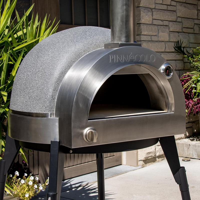 The Fire One Up PINNACOLO L’ARGILLA Gas Fired Outdoor Clay Pizza Oven with Accessories (PPO-8-08) features a rounded stainless steel top, chimney, and sturdy black legs, set on a patio surrounded by greenery and stone walls.