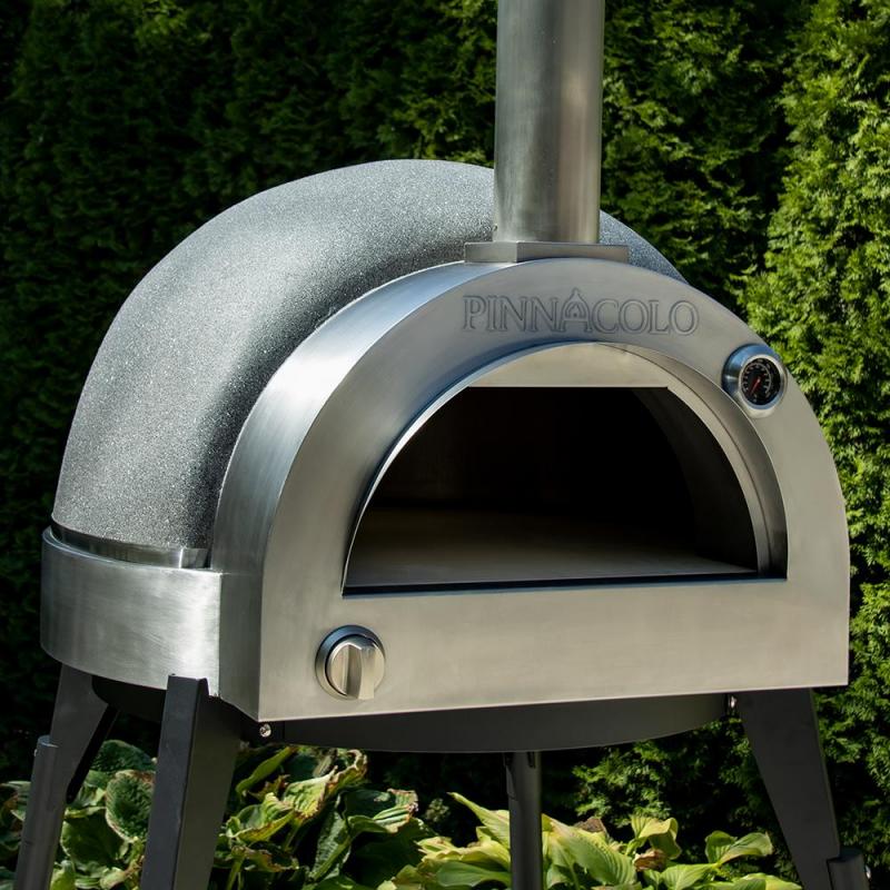 The Fire One Up PINNACOLO L’ARGILLA Gas Fired Outdoor Clay Pizza Oven with Accessories (PPO-8-08) features a domed top and chimney, and stands on black legs in a garden surrounded by green shrubs.