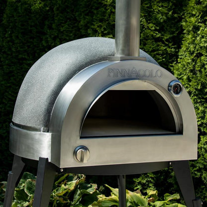 The Fire One Up PINNACOLO L’ARGILLA Gas Fired Outdoor Clay Pizza Oven with Accessories (PPO-8-08) features a domed top and chimney, and stands on black legs in a garden surrounded by green shrubs.