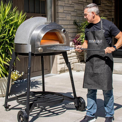 A man in a black apron stands beside the Fire One Up PINNACOLO L’ARGILLA Gas Fired Outdoor Clay Pizza Oven (PPO-8-08) on wheels, holding a pizza peel. The dome-shaped oven is on a patio with plants and a stone wall behind.