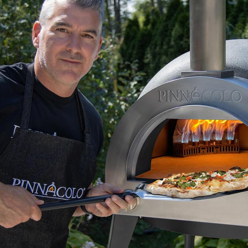 A man in a black PINNACOLO apron removes a fresh pizza using a peel from the Fire One Up PINNACOLO L’ARGILLA Gas Fired Outdoor Clay Pizza Oven (PPO-8-08), with visible flames inside.