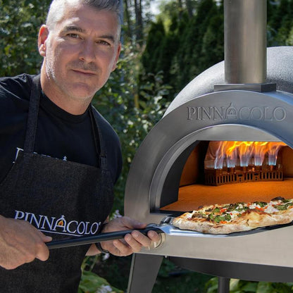 A man in a black PINNACOLO apron removes a fresh pizza using a peel from the Fire One Up PINNACOLO L’ARGILLA Gas Fired Outdoor Clay Pizza Oven (PPO-8-08), with visible flames inside.