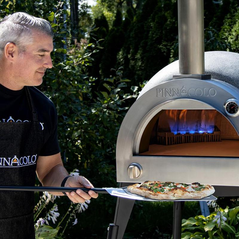 A man uses a pizza peel to place a freshly baked pizza into the Fire One Up PINNACOLO L’ARGILLA Gas Fired Outdoor Clay Pizza Oven (PPO-8-08) with visible blue flame, surrounded by greenery.