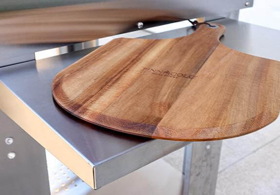 Side shelf of the PINNACOLO PREMIO Wood-Fired Outdoor Pizza Oven with a branded 'PINNACOLO' wooden peel placed on top.