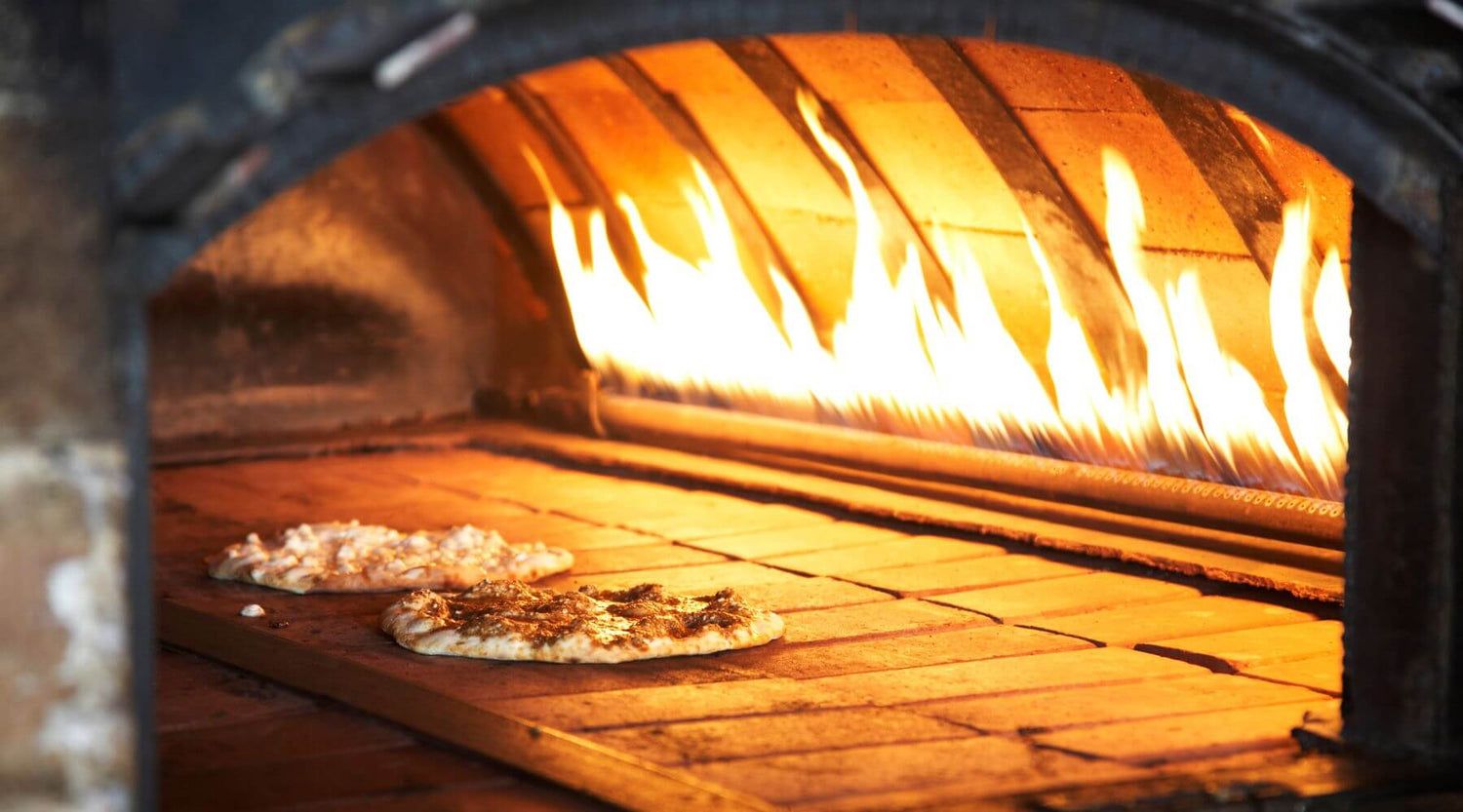 Two pizzas with different toppings bake on the stone surface of a large gas-fired oven, with tall flames shooting upward in the background.
