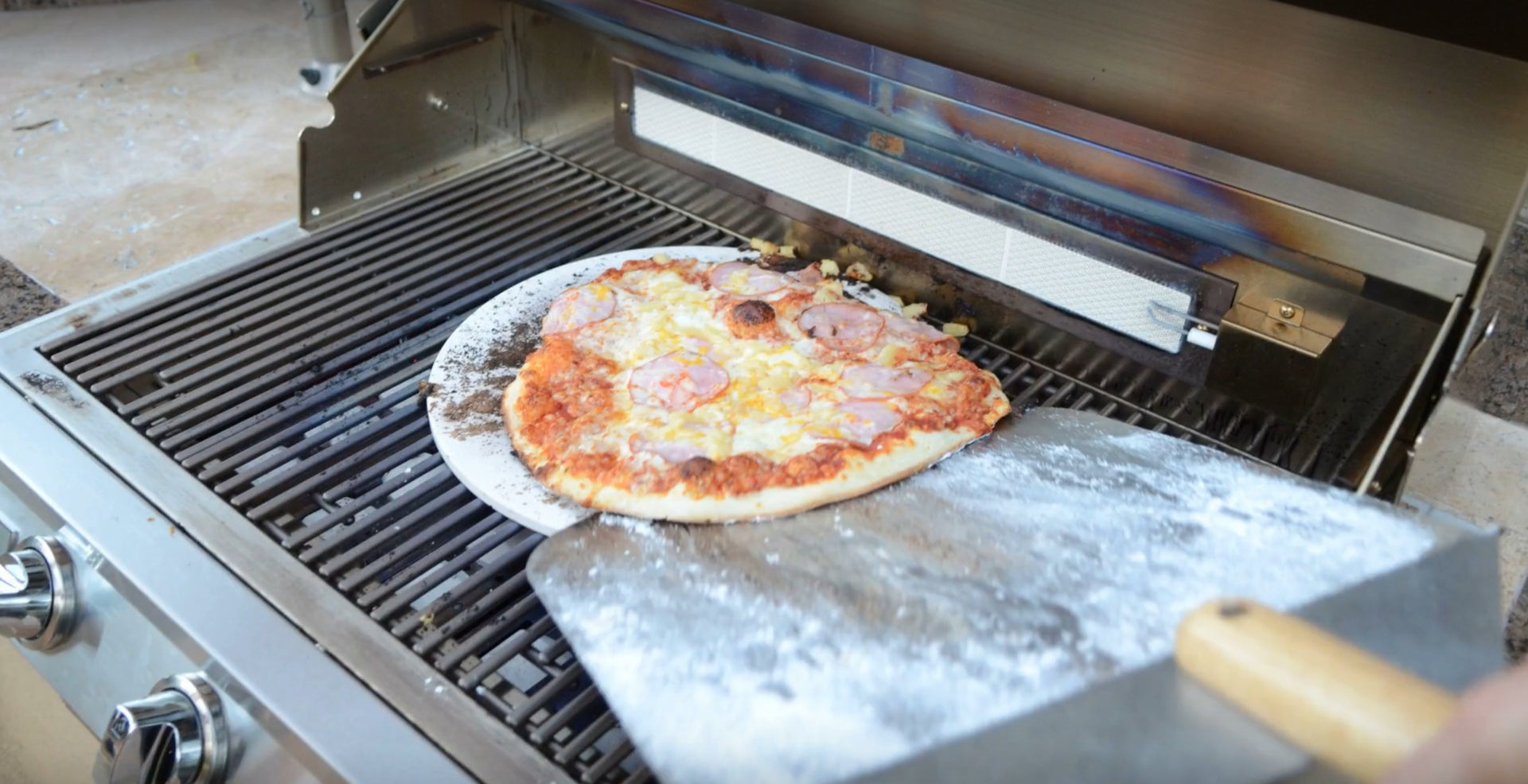 A pizza on the Kokomo Grills 14-Inch Pizza Stone Kamado Ceramic Oven Insert is being removed from the grill with a metal peel, cheese melted perfectly and flour visible—just like a wood-fired oven.