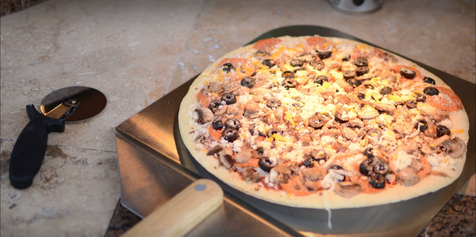 A wood-fired pizza topped with mushrooms, black olives, and cheese rests on a Kokomo Grills 14-Inch Pizza Stone Kamado Ceramic Oven Insert by Kokomo Grills, with a pizza cutter beside it on the stone countertop.