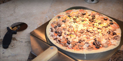 A wood-fired pizza topped with mushrooms, black olives, and cheese rests on a Kokomo Grills 14-Inch Pizza Stone Kamado Ceramic Oven Insert by Kokomo Grills, with a pizza cutter beside it on the stone countertop.