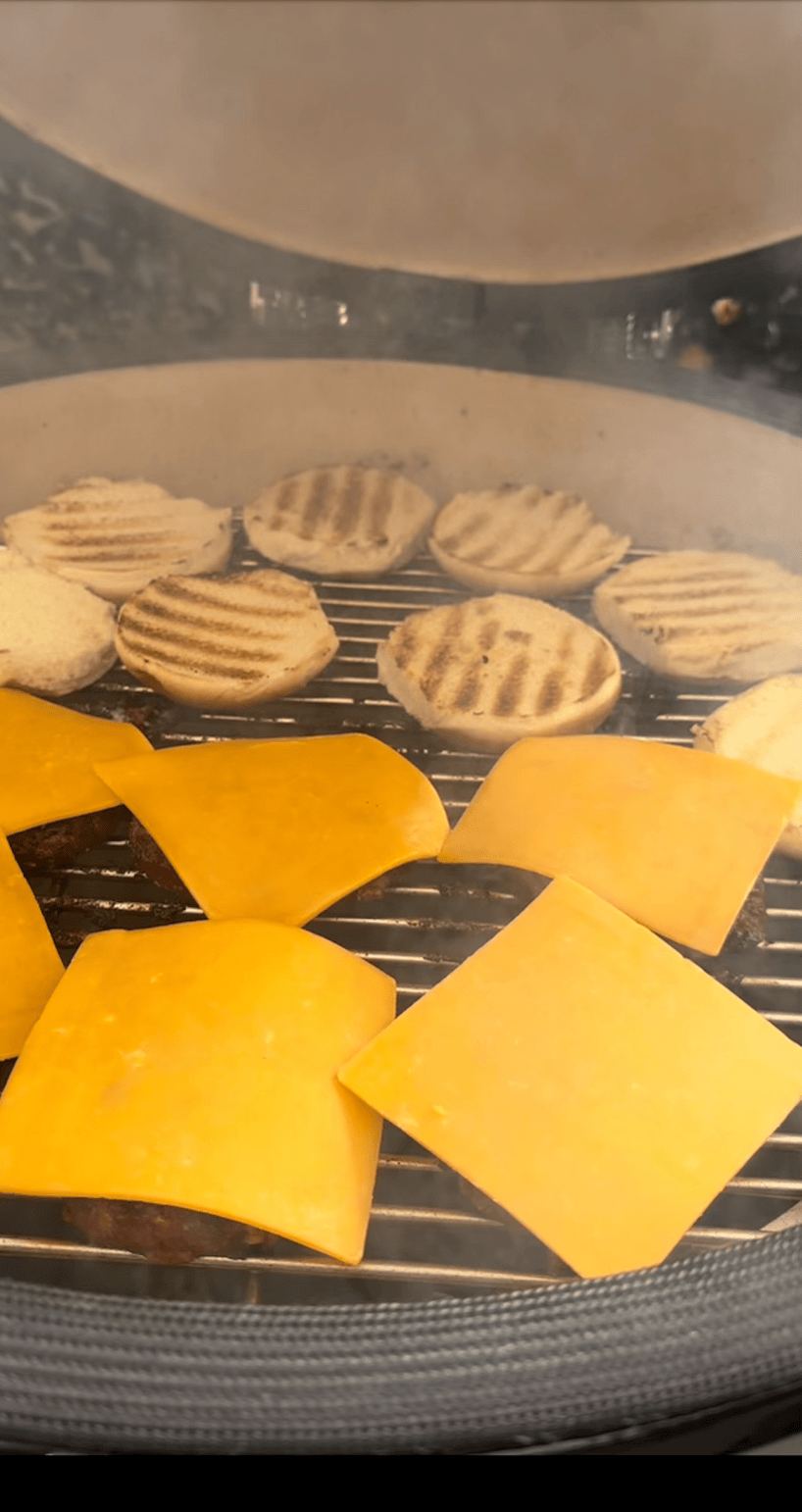 Sliced cheddar cheese melts over burger patties as halved, toasted buns grill inside the Kokomo Grills 21-Inch Kamado Ceramic BBQ Grill KO-21-KAMADO, with the lid partially open for smoky, charcoal-infused flavor.