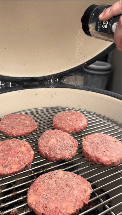 Six raw hamburger patties sizzle on the Kokomo Grills 21-Inch Kamado Ceramic BBQ Grill KO-21-KAMADO as a hand sprinkles seasoning from a shaker, infusing smoky flavor into classic charcoal grilling.