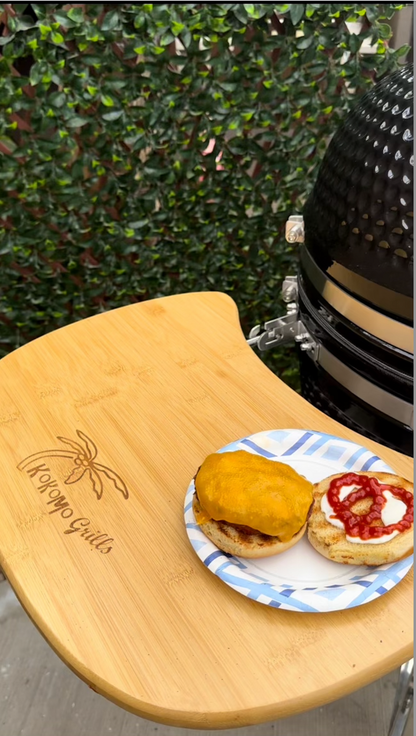 A bamboo grill side table holds a paper plate with two burger patties—one with melted cheese, one with ketchup and mayonnaise. The Kokomo Grills 21-Inch Kamado Ceramic BBQ Grill KO-21-KAMADO sits nearby amid green leaves, perfect for charcoal cooking.