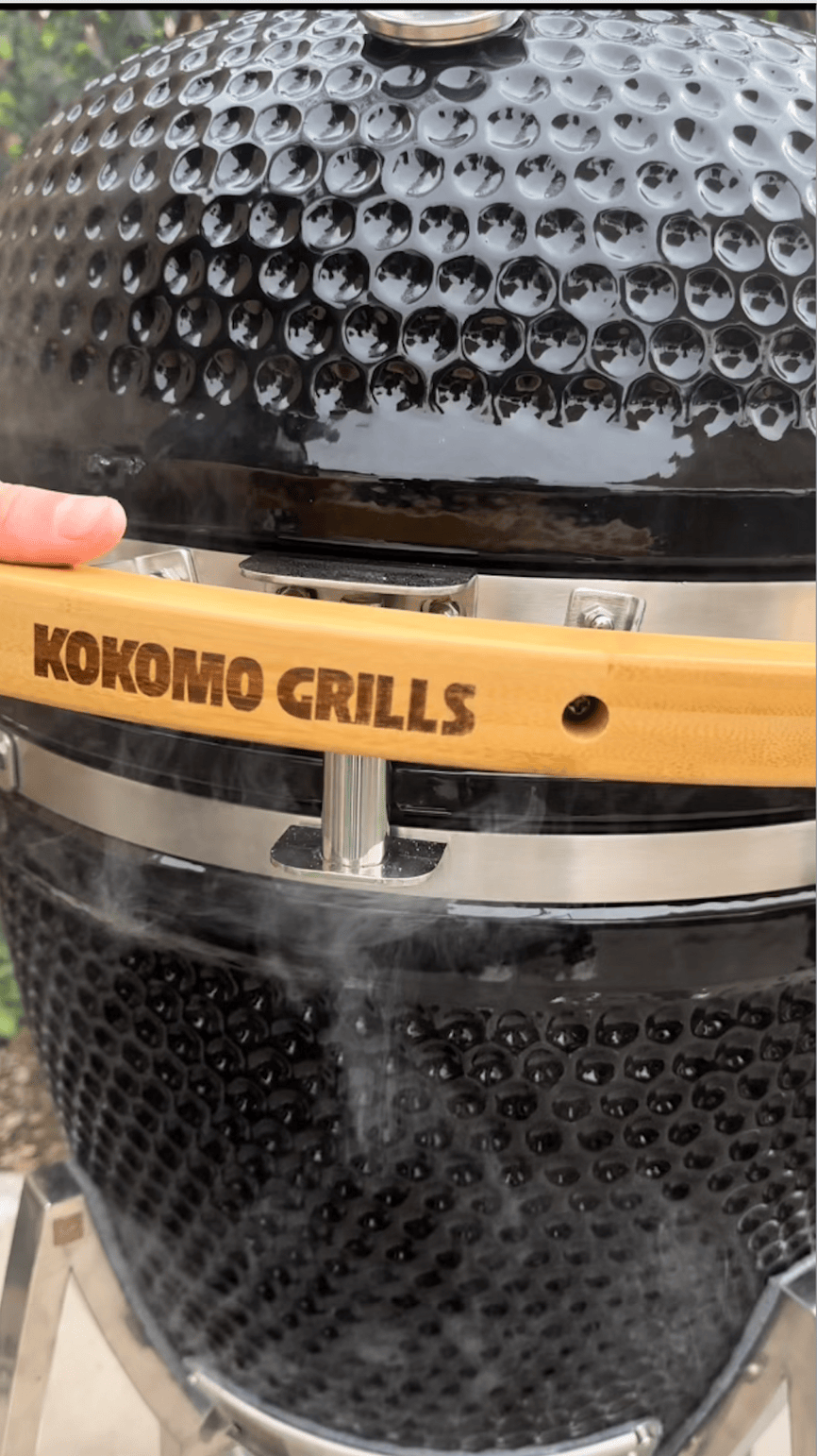 A close-up of the Kokomo Grills 21-Inch Kamado Ceramic BBQ Grill KO-21-KAMADO shows smoke rising as a hand touches its wooden handle, capturing the essence of charcoal cooking with this premium grill from Kokomo Grills.