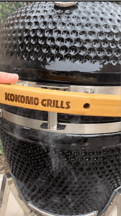 A close-up of the Kokomo Grills 21-Inch Kamado Ceramic BBQ Grill KO-21-KAMADO shows smoke rising as a hand touches its wooden handle, capturing the essence of charcoal cooking with this premium grill from Kokomo Grills.