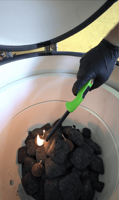 A person wearing a black glove uses a green lighter to ignite a fire starter among charcoal briquettes inside the Kokomo Grills 21-Inch Kamado Ceramic BBQ Grill KO-21-KAMADO, ideal for authentic charcoal grilling.