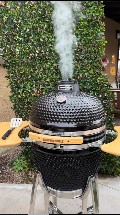 The Kokomo Grills 21-Inch Kamado Ceramic BBQ Grill KO-21-KAMADO stands on a metal frame, smoke rising from its top vent. Utensils rest on wooden side shelves, backed by a green hedge—perfect for authentic charcoal grilling.