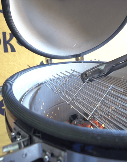 A close-up of the Kokomo Grills 21-Inch Kamado Ceramic BBQ Grill KO-21-KAMADO shows its open lid, with a metal grate being lifted by tongs and sparks rising from burning charcoal. A yellow banner appears in the background.