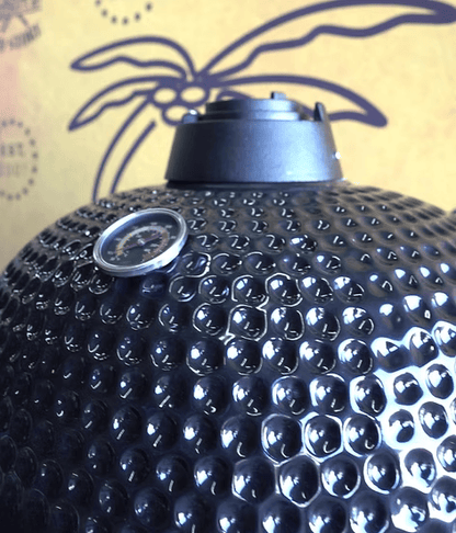 A close-up of the Kokomo Grills 21-Inch Kamado Ceramic BBQ Grill (KO-21-KAMADO) in black features a dimpled finish, built-in thermometer, and top vent. A yellow wall with a palm tree design is seen in the background—ideal for charcoal grill fans.