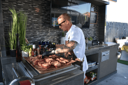 A man in sunglasses and gloves grills meat on a Kokomo Grills 32-Inch Built-In Charcoal Grill in his outdoor kitchen, surrounded by cooking supplies and greenery, with a "No Singing, No Running, No Dancing" sign behind him.