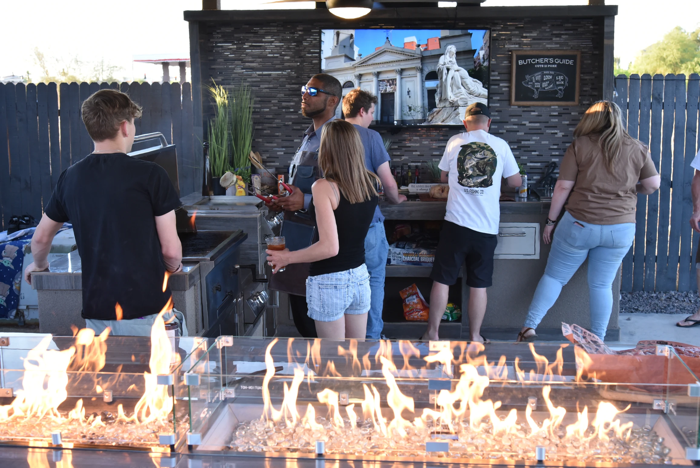 Five people enjoy cooking together at a Kokomo Grills 32-Inch Built-In Charcoal Grill in a festive, casual outdoor kitchen. A fire pit glows in front, while a mural and butcher’s guide decorate the wall behind them.