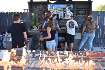 Five people enjoy cooking together at a Kokomo Grills 32-Inch Built-In Charcoal Grill in a festive, casual outdoor kitchen. A fire pit glows in front, while a mural and butcher’s guide decorate the wall behind them.
