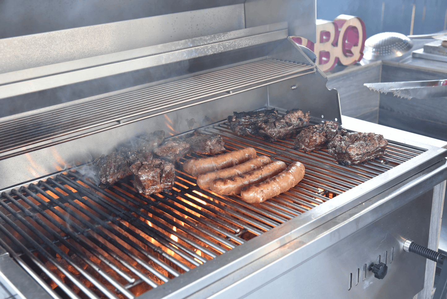 Several sausages and steaks sizzle on the Kokomo Grills 32-Inch Built-In Charcoal Grill, flames below and smoke rising, as BBQ-themed decorations create an inviting outdoor kitchen scene.