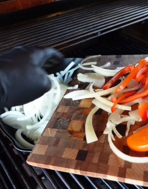 A gloved hand slides sliced onions and red bell peppers from a wooden cutting board onto the Kokomo Grills 32-Inch Built-In Charcoal Grill, prepping fresh veggies for an outdoor feast.