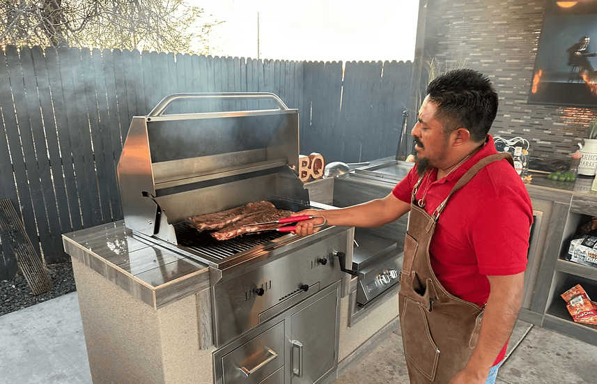 A man in a red shirt and brown apron grills ribs with tongs on a Kokomo Grills 32-Inch Built-In Charcoal Grill, made of 304 stainless steel, as smoke rises in his modern outdoor kitchen patio.