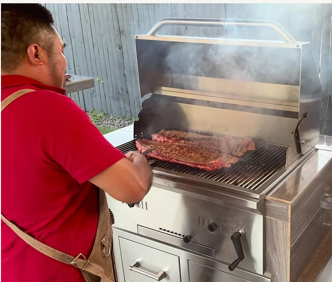 Wearing a red shirt, a person cooks a large piece of meat on the Kokomo Grills 32-Inch Built-In Charcoal Grill in an outdoor kitchen, smoke rising beside a wooden fence.