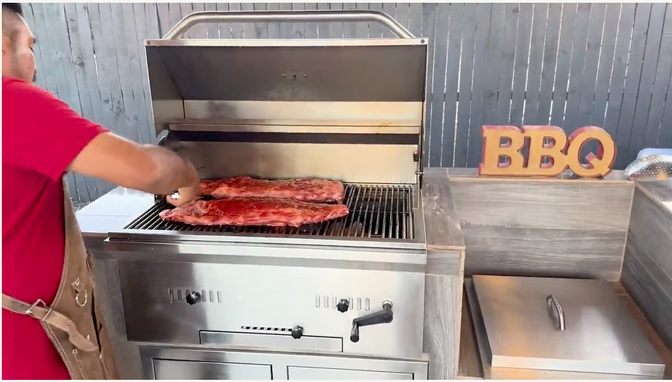 Wearing a red shirt, someone grills two racks of ribs on a Kokomo Grills 32-Inch Built-In Charcoal Grill, made from 304 stainless steel, with a decorative BBQ sign nearby in their stylish outdoor kitchen.