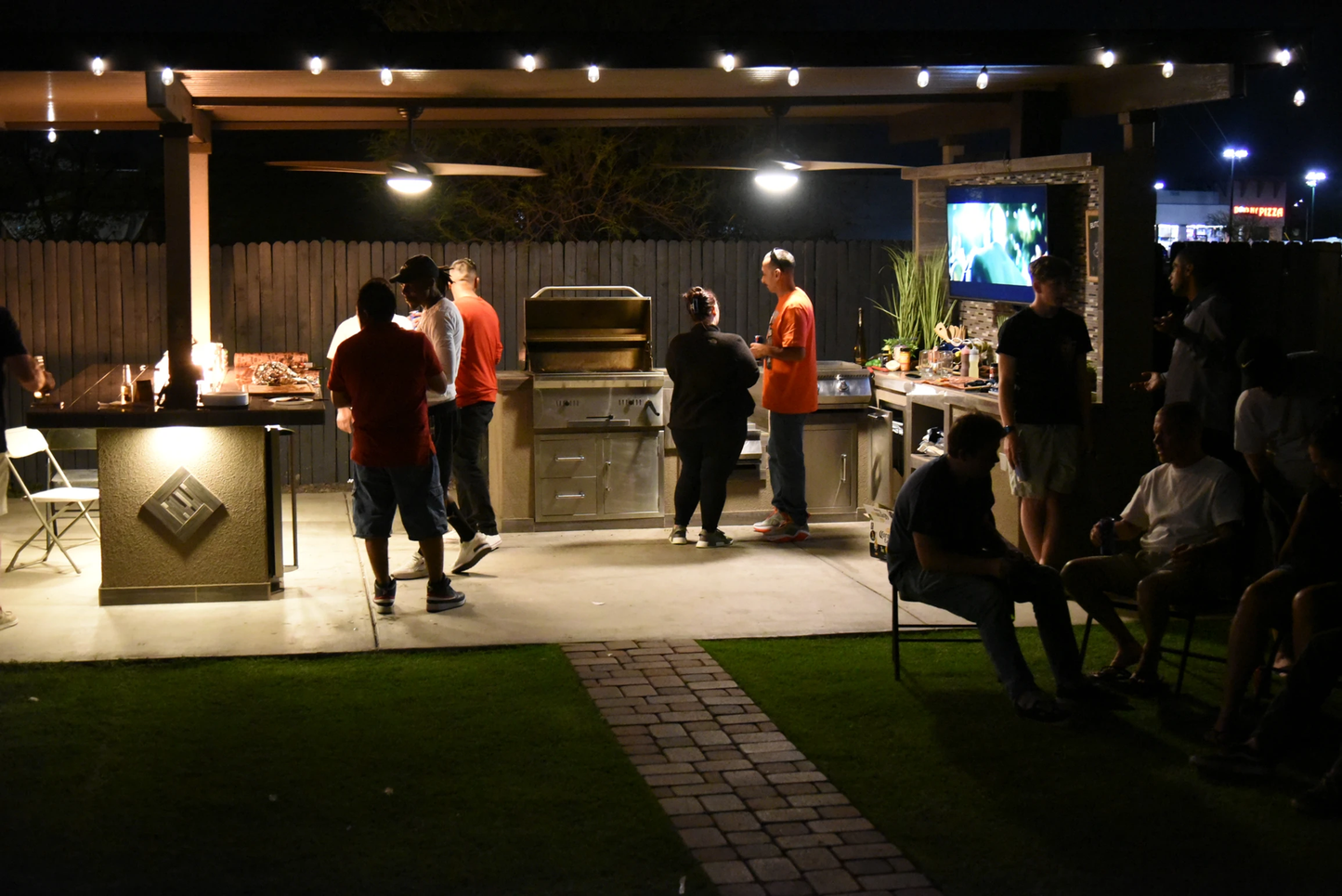 At night, people gather on a patio under string lights, some grilling with the Kokomo Grills 32-Inch Built-In Charcoal Grill, while others sit nearby chatting and enjoying a relaxed barbecue.