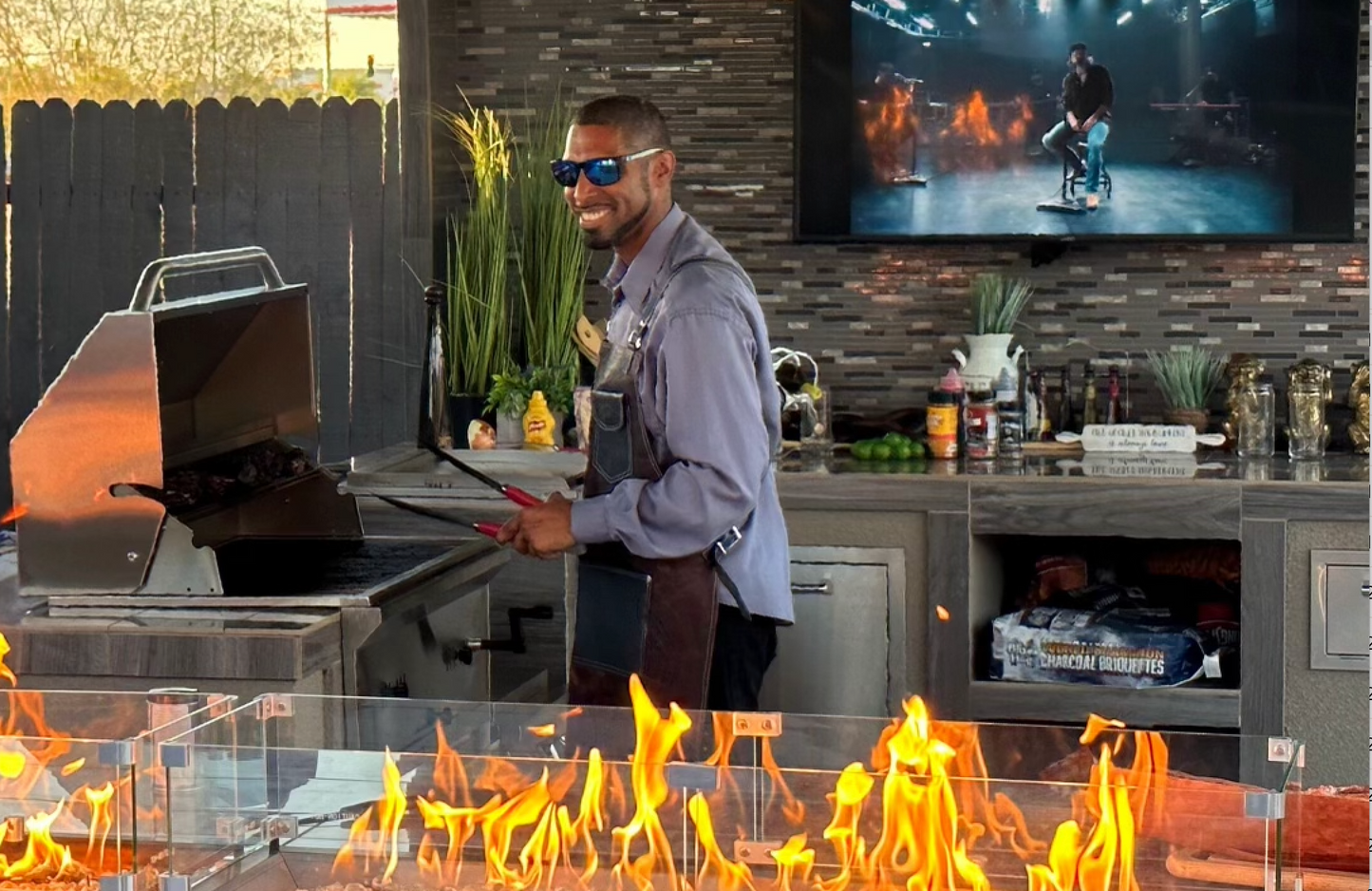 A man in sunglasses smiles while grilling on a Kokomo Grills 32-Inch Built-In Charcoal Grill outdoors by a bright fire; the kitchen features spices and ingredients, with a TV showing a music performance with fire effects in the background.