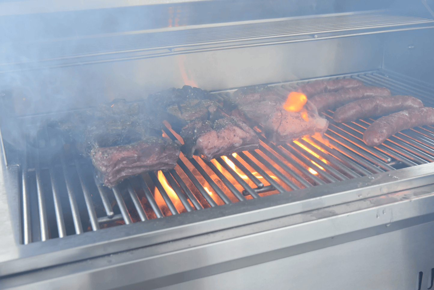 Several cuts of meat and sausages cook over visible flames and smoke on the Kokomo Grills 32-Inch Built-In Charcoal Grill, creating a lively barbecue scene in an outdoor kitchen.
