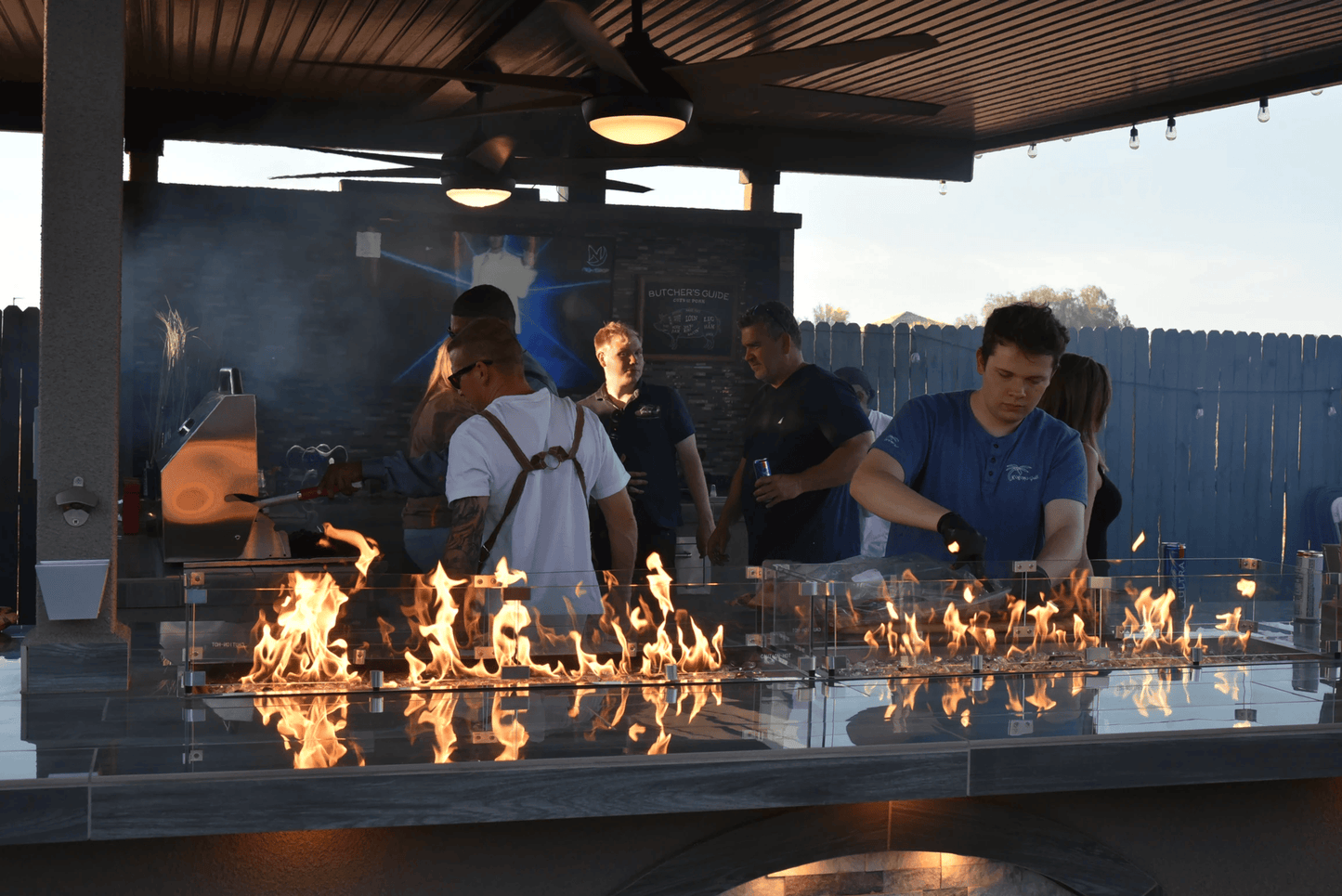 Friends cook and chat around a Kokomo Grills 32-Inch Built-In Charcoal Grill, enjoying its warmth under a covered patio as evening falls.